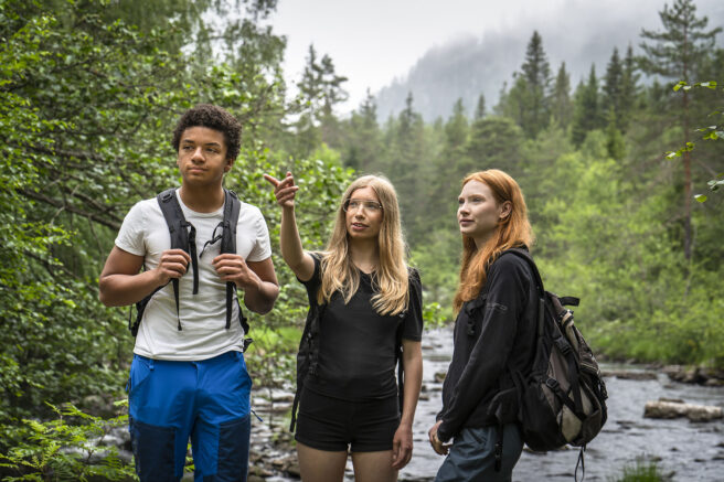 Tre unge mennesker i skogen. En elv og grønne trær i bakgrunnen. De har på seg turklær. En av dem peker på noe utenfor bildet og alle tre ser i den retningen.