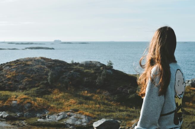 En ung person med langt, brunt hår står med ryggen til i et norsk naturlandskap og ser ut over havet.